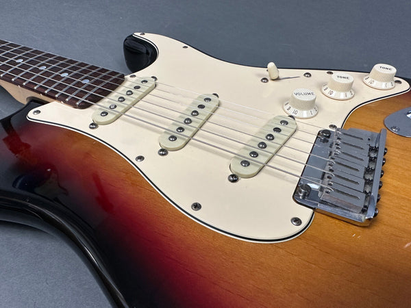 Close-up of sunburst electric guitar body with white pickguard, three single-coil pickups, volume and tone control knobs, and chrome bridge on a gray background