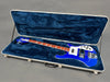 Blue electric bass guitar with white pickguard in hard shell case lined with dark blue plush interior on gray floor