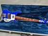Blue Rickenbacker electric bass guitar with white pickguard and triangular inlays, displayed in blue plush-lined hardshell case
