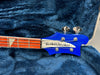 Close-up of Rickenbacker bass guitar headstock in blue with silver tuning pegs and fretboard inlay, displayed in a plush blue guitar case interior.