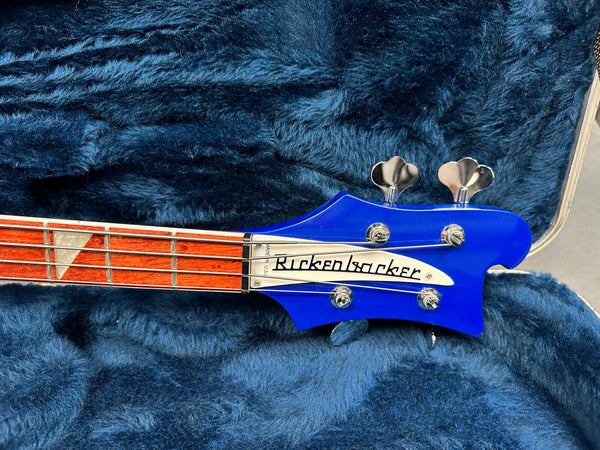 Close-up of Rickenbacker bass guitar headstock in blue with silver tuning pegs and fretboard inlay, displayed in a plush blue guitar case interior.