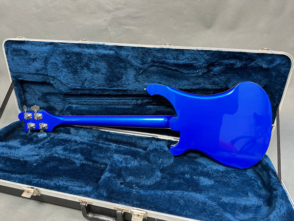 Electric bass guitar with blue finish inside blue plush-lined hard case showing the back of the instrument and tuning pegs