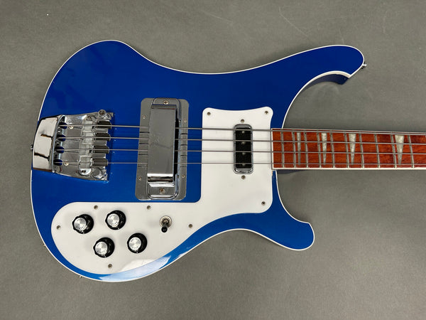 Blue electric bass guitar with white pickguard and chrome bridge hardware, wooden fretboard with triangular inlays, four control knobs, and toggle switch on gray background