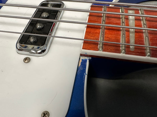 Close-up of electric bass guitar strings, chrome pickup, white pickguard, and rosewood fretboard with inlay, showing wear on blue guitar body edge.