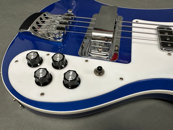 Close-up of blue electric bass guitar body showing chrome bridge, pickups, black tone and volume control knobs on white pickguard, and toggle switch