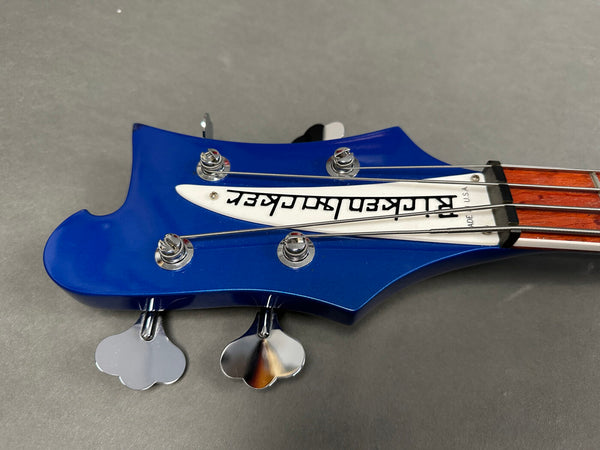 Close-up of blue Rickenbacker bass guitar headstock with chrome tuning pegs and wood fretboard against gray background