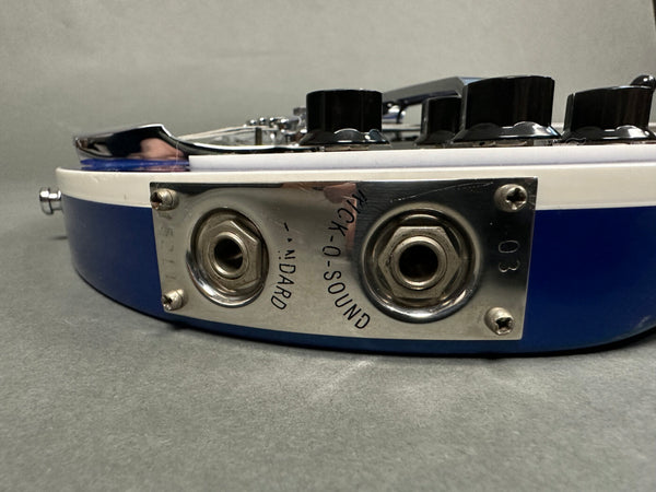 Close-up of blue electric guitar input jack panel with two output ports labeled "Standard" and "Mick-O-Sound" and volume control knobs visible on the body