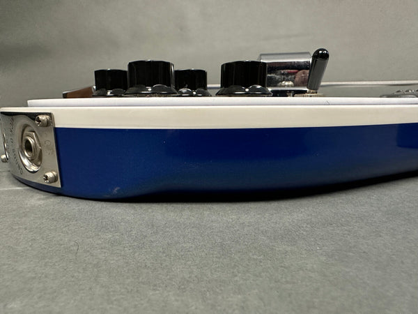 Close-up side view of blue and white electric guitar body with black control knobs and input jack plate on gray background