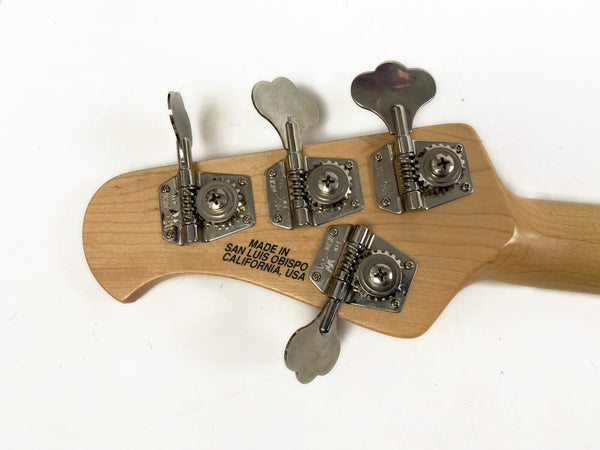 Close-up of bass guitar headstock with four metal tuning pegs and "Made in San Luis Obispo California, USA" printed on natural wood finish