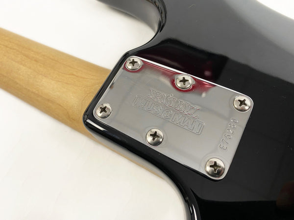Close-up of Ernie Ball Music Man guitar neck plate with serial number E775280 on a black electric guitar body and natural wood neck.