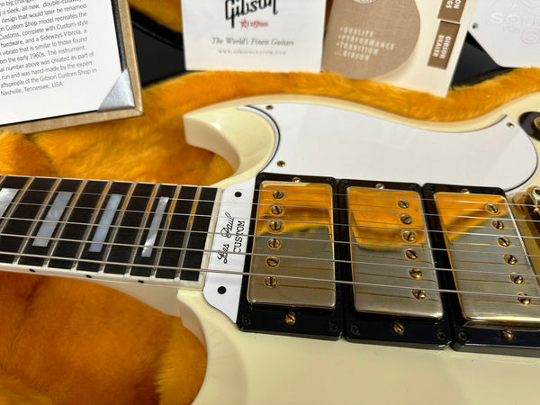 Close-up of cream Gibson Les Paul Custom electric guitar body and neck showing triple gold humbucker pickups and pearl block inlays on ebony fretboard, displayed in orange plush-lined guitar case with Gibson Custom Shop certification papers.