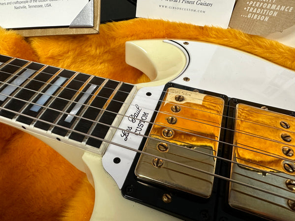 Close-up of a vintage white Gibson Les Paul Custom electric guitar showing the fretboard with block inlays and gold-plated humbucker pickups, resting on a plush orange guitar case interior.