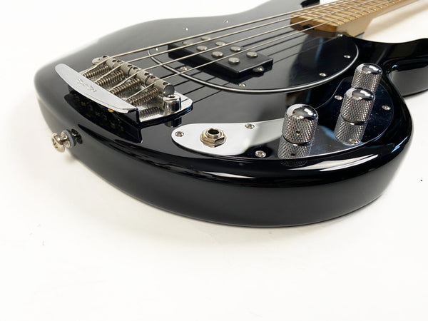 Close-up of black electric bass guitar body showing chrome bridge, control knobs, pickup, and output jack on white background