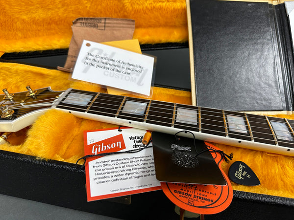 Close-up of Gibson Custom guitar neck and fretboard inside yellow plush-lined hardshell case with Gibson vintage tone circuit card, Certificate of Authenticity, Gibson Custom hang tag, and black Gibson Pure guitar pick.