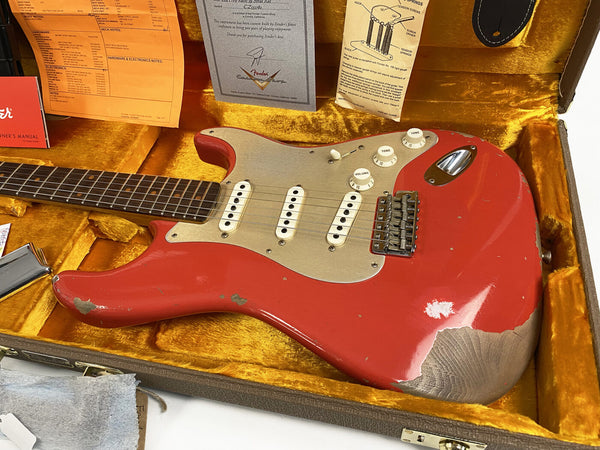 Vintage red Fender Stratocaster electric guitar with worn paint finish, three single-coil pickups, white control knobs, and rosewood fretboard inside a plush gold-lined guitar case with Fender manuals and documentation.