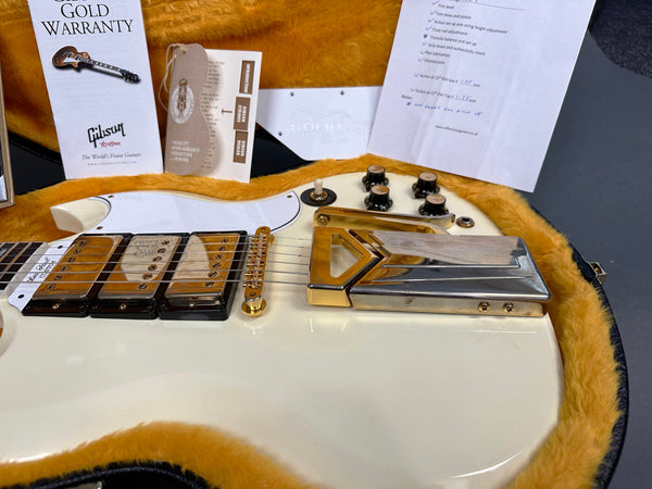 Close-up of white electric guitar with gold hardware, three gold pickups, and black control knobs in yellow fur-lined guitar case, accompanied by Gibson Gold Warranty and setup documents.