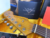Close-up of Fender Stratocaster guitar headstock with vintage tuning pegs and strings, displayed in yellow plush-lined guitar case with Fender Custom Shop Custom Care booklet and other accessories.