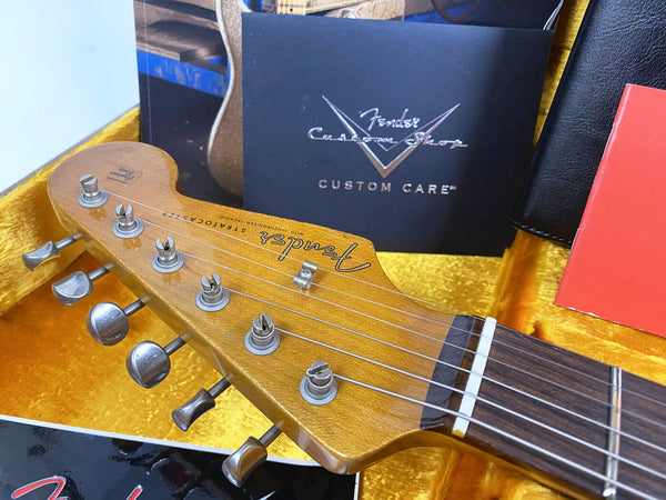Close-up of Fender Stratocaster guitar headstock with vintage tuning pegs and strings, displayed in yellow plush-lined guitar case with Fender Custom Shop Custom Care booklet and other accessories.