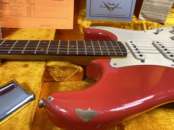 Close-up of a vintage red Fender Stratocaster electric guitar with worn paint and a rosewood fretboard, resting in a yellow plush guitar case with Fender Custom Shop warranty and notes visible.