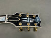 Close-up of Gibson guitar headstock with black finish, gold tuning pegs, and mother-of-pearl inlay on gray background
