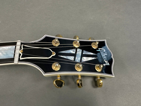 Close-up of Gibson guitar headstock with black finish, gold tuning pegs, and mother-of-pearl inlay on gray background