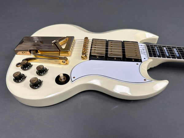 Close-up of white electric guitar body with gold hardware, triple humbucker pickups, vibrato tailpiece, white pickguard, and black control knobs on gray background.