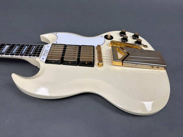 White electric guitar with gold hardware, triple humbucker pickups, white pickguard, and four black control knobs on gray background