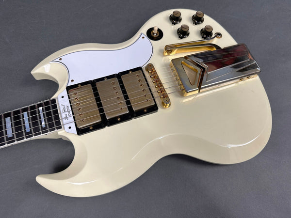 Vintage white electric guitar close-up showing three gold humbucker pickups, white pickguard, gold bridge and vibrato tailpiece, black control knobs on gray background