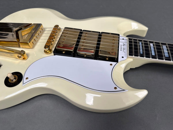 Close-up of white electric guitar body with three gold humbucker pickups, white pickguard, gold bridge, and black and white toggle switch on gray background