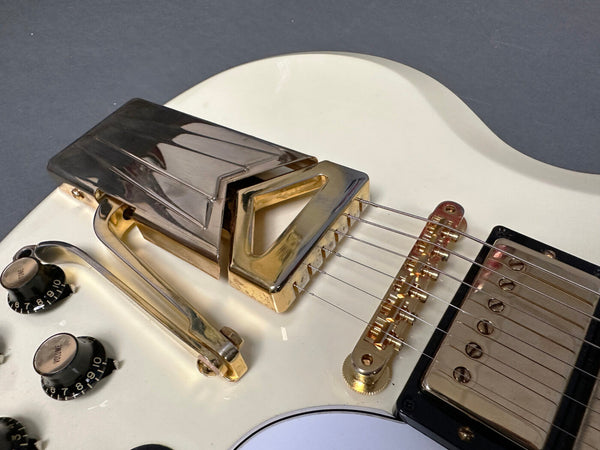 Close-up of white electric guitar body showing gold hardware including vibrato tailpiece, bridge, volume and tone knobs, and humbucker pickup