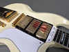 Close-up of white electric guitar body with gold hardware, triple humbucker pickups, and white pickguard with "Les Paul Custom" label near the neck.