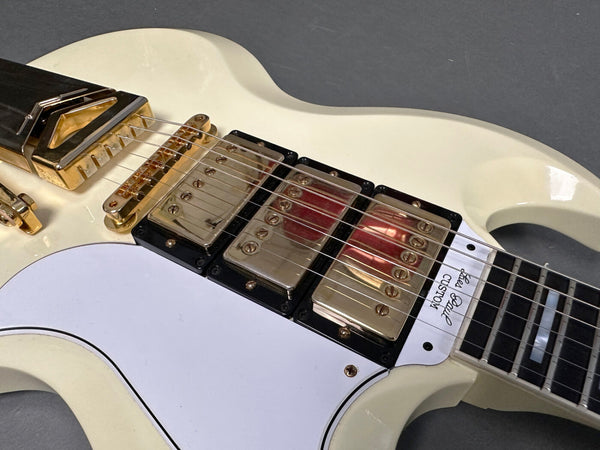 Close-up of white electric guitar body with gold hardware, triple humbucker pickups, and white pickguard with "Les Paul Custom" label near the neck.