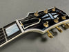 Close-up of Gibson guitar headstock with black finish, mother-of-pearl inlay, and gold tuning pegs on gray background