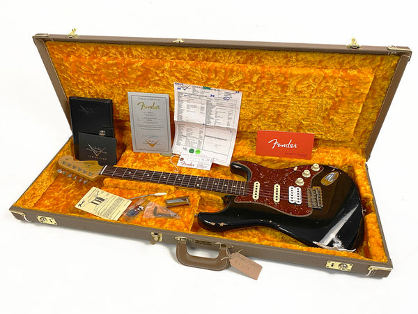 Vintage black Fender Stratocaster electric guitar with tortoiseshell pickguard in open brown hard case with orange plush interior, including Fender owner's manual, certificate of authenticity, custom care booklet, paperwork, and guitar accessories