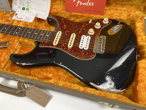 Vintage Fender Stratocaster electric guitar with black finish, tortoiseshell pickguard, humbucker pickup, and three control knobs, displayed in orange plush-lined case with Fender owner's manual and authenticity tags.