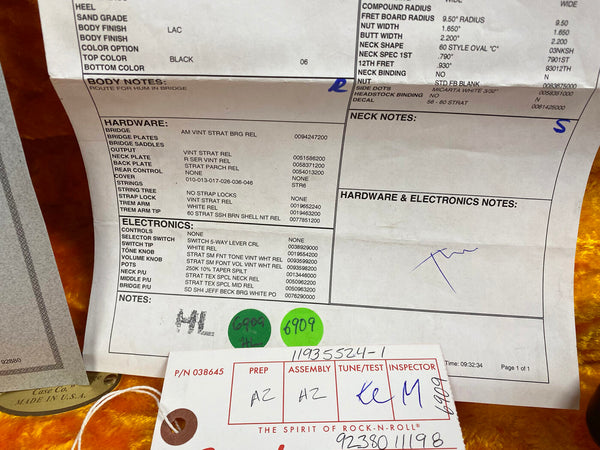 Close-up of vintage style Stratocaster guitar build specifications sheet detailing hardware, electronics, and assembly notes with part numbers and inspector signatures, on an orange guitar case background.
