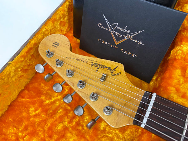 Close-up of Fender Stratocaster guitar headstock with tuning pegs, maple wood finish, and black Fender Custom Shop Custom Care card on orange plush interior guitar case lining