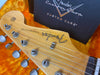 Close-up of Fender Stratocaster guitar headstock with tuning pegs and string tree, resting on orange plush guitar case interior with Fender Custom Shop Custom Care box in background