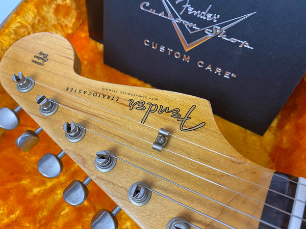 Close-up of Fender Stratocaster guitar headstock with tuning pegs and string tree, resting on orange plush guitar case interior with Fender Custom Shop Custom Care box in background