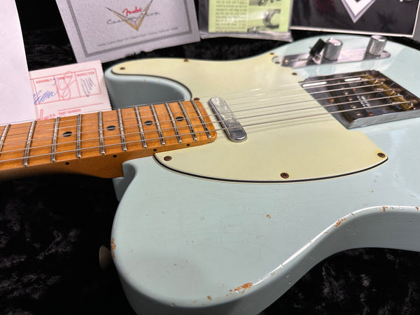 Close-up of vintage Fender Telecaster electric guitar with light blue finish, maple neck, and aged pickguard showing wear and patina, photographed on black plush surface with Fender certificate and paperwork in background.