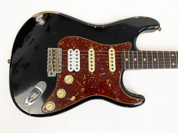 Black electric guitar with tortoiseshell pickguard and white humbucker and single-coil pickups, worn vintage style with visible paint chips and scratches, close-up view of body and neck on white background.