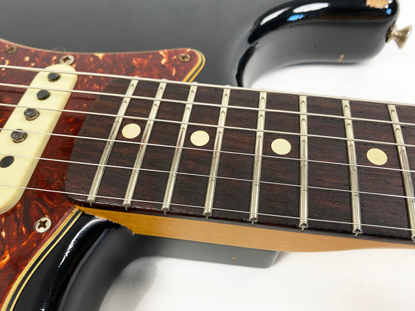Close-up of electric guitar neck and fretboard with dot inlays, tortoiseshell pickguard, and black body finish showing worn edges and metal strings.