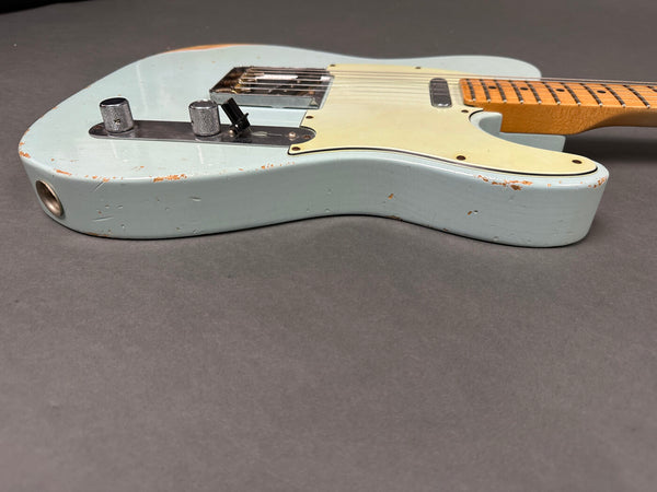 Light blue vintage-style electric guitar with a maple neck and aged finish showing paint wear on the body, positioned on a gray surface.