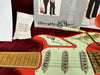 Close-up of Burns London electric guitar body and maple neck with vintage-style pickups, cream pickguard, gold hardware, and a vibrato arm in plush red guitar case with silica gel packet and signed certificate in the background.