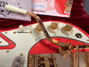 Close-up of red Burns electric guitar with gold tremolo arm, white tone and volume knobs, and vintage Burns London promotional materials in background