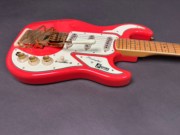 Close-up of red Burns London electric guitar with white pickguard, three single-coil pickups, gold Bigsby-style tremolo, and maple neck on gray background