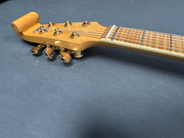 Close-up of a natural wood electric guitar headstock with gold tuning pegs and steel strings on a gray background
