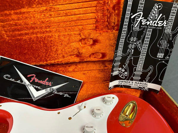 Close-up of red Fender electric guitar body with white pickguard, tone and volume knobs, gold input jack, Fender Custom Shop certificate, and Fender owner's manual inside orange plush guitar case interior.