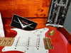 Close-up of red Fender Stratocaster electric guitar with white pickguard, three single-coil pickups, white tone and volume knobs, gold bridge, and Fender owner's manual inside orange velvet guitar case