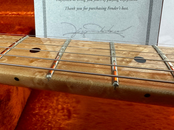 Close-up of a maple guitar neck with dot inlays and metal frets, showing guitar strings and part of a certificate in the background inside an orange padded guitar case.